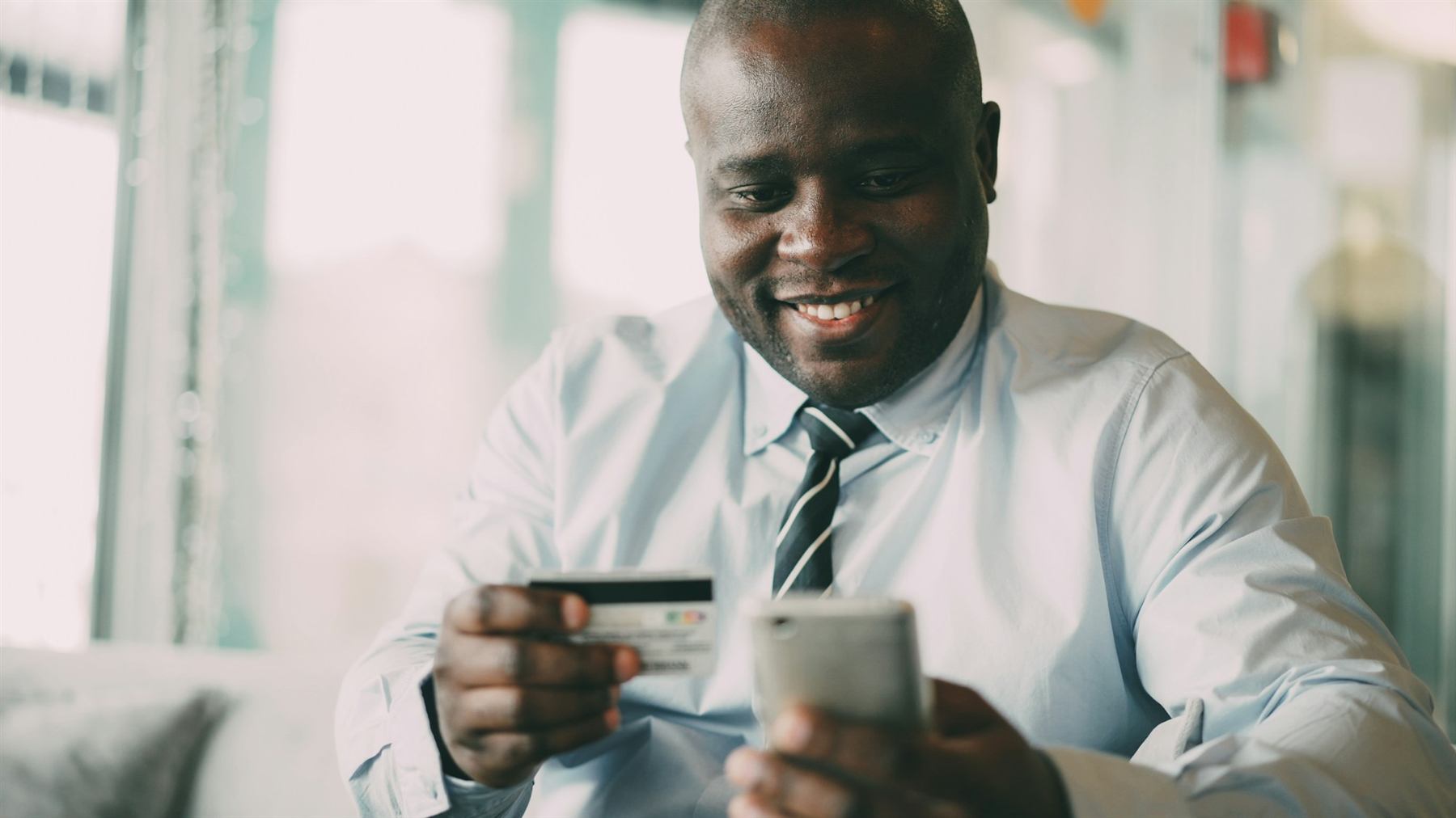 A smiling man in a café holding a bank card in one hand and his phone in the other
